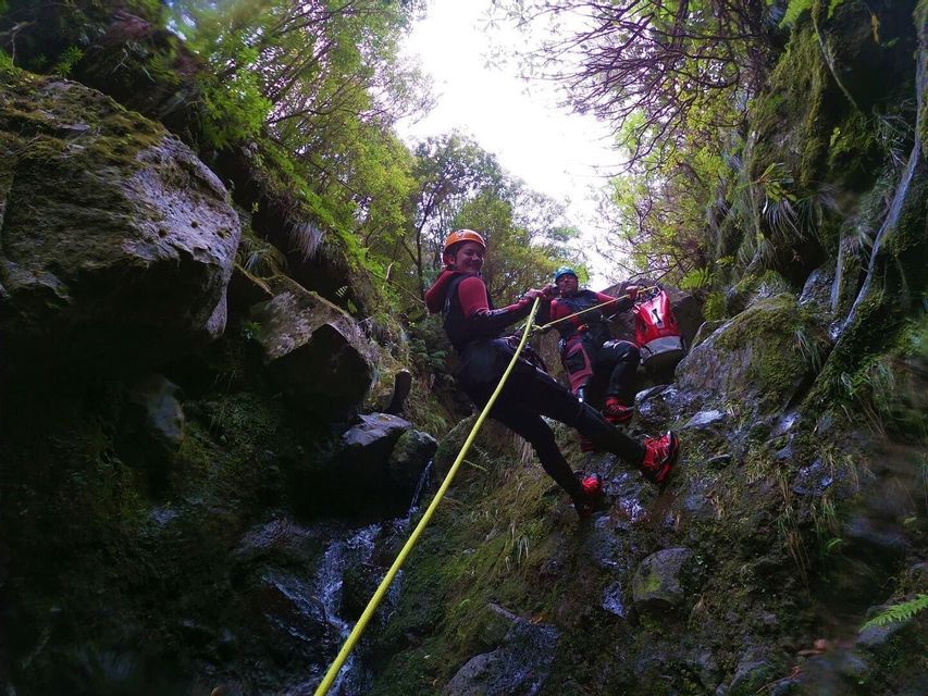 Dos personas de un viaje en grupo de WeRoad, con cascos y trajes de neopreno, haciendo rápel por una pared rocosa musgosa en un cañón.