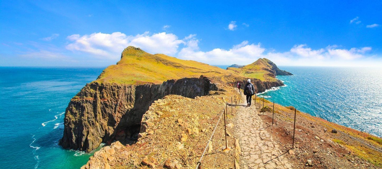 Una persona con mochila haciendo senderismo por un estrecho sendero de piedra junto a un alto acantilado costero cubierto de hierba con vistas al océano.