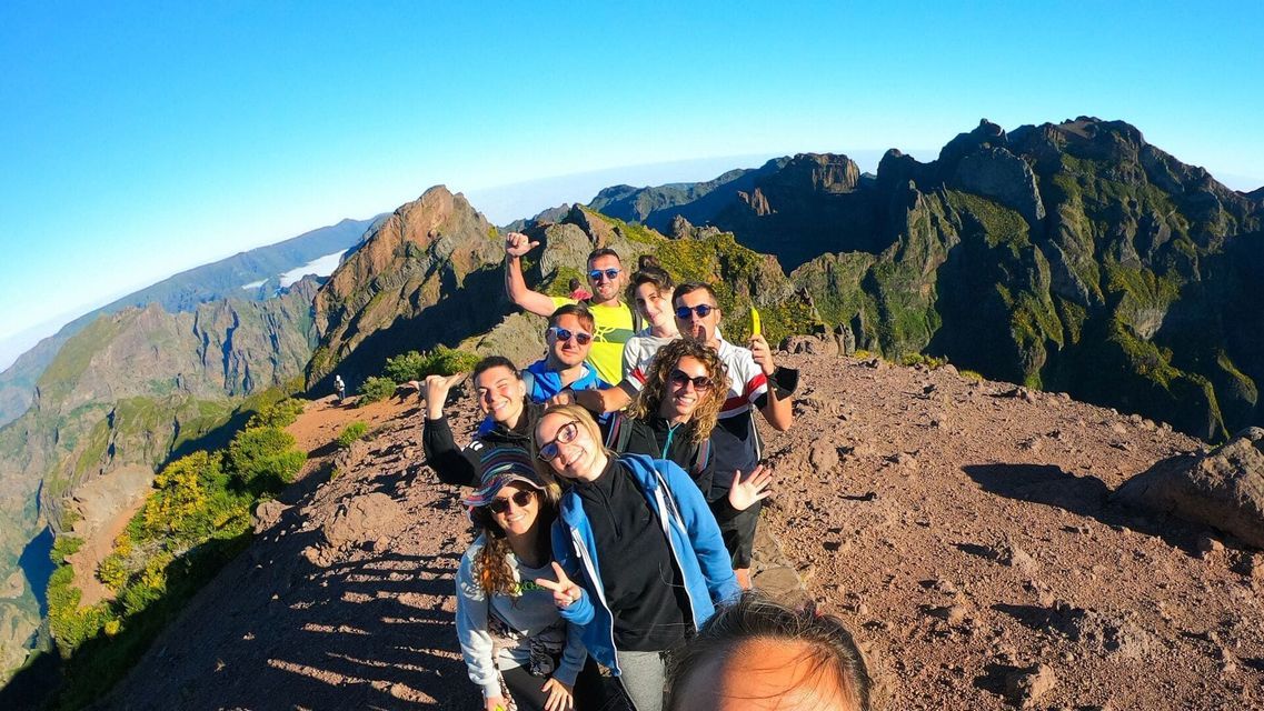 A WeRoad group trip takes a selfie on a sunny mountain peak, with a rugged green mountain range visible in the background.