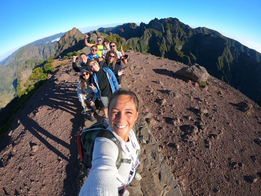 A woman takes a smiling selfie with her WeRoad group trip on a narrow, rocky mountain ridge under a clear blue sky.