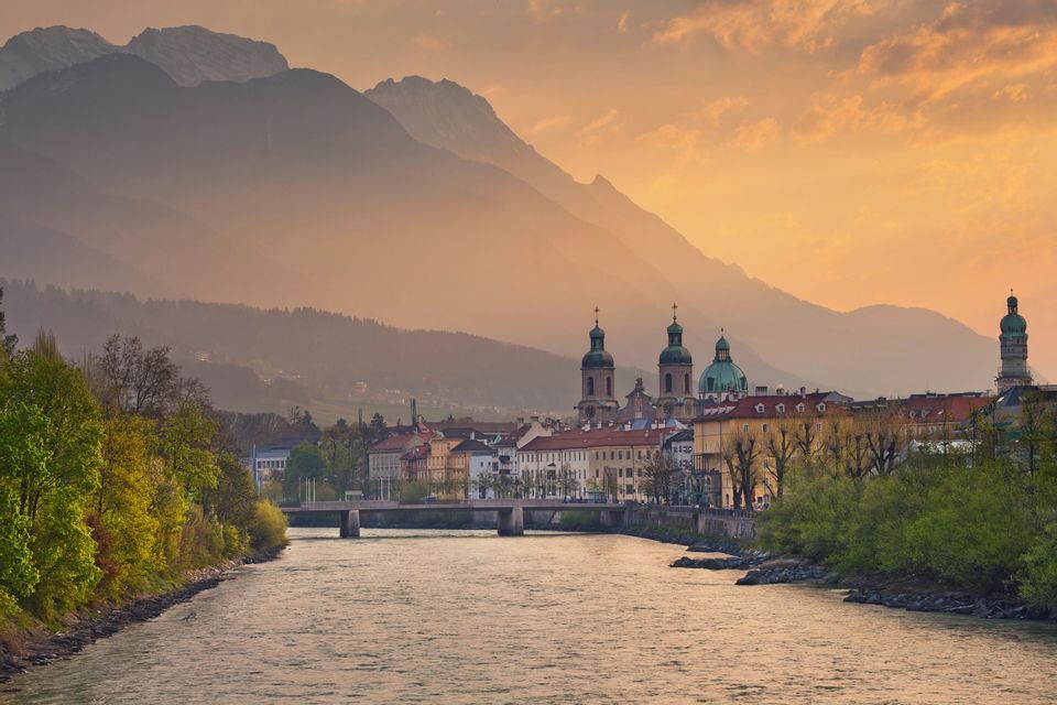 Ein breiter Fluss fließt an einer historischen Stadt mit grünkuppeligen Kirchtürmen vorbei, vor der Kulisse dunstiger Berge im Sonnenuntergang.