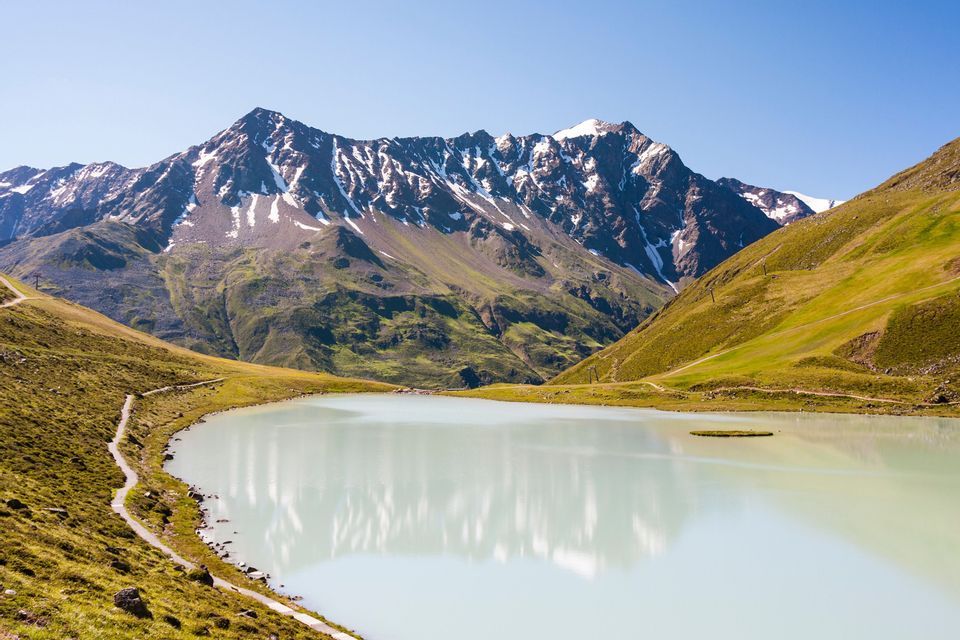Ein milchig-blauer Alpensee spiegelt eine schneebedeckte Bergkette wider, mit einem gewundenen Pfad entlang seines grasbewachsenen Ufers.