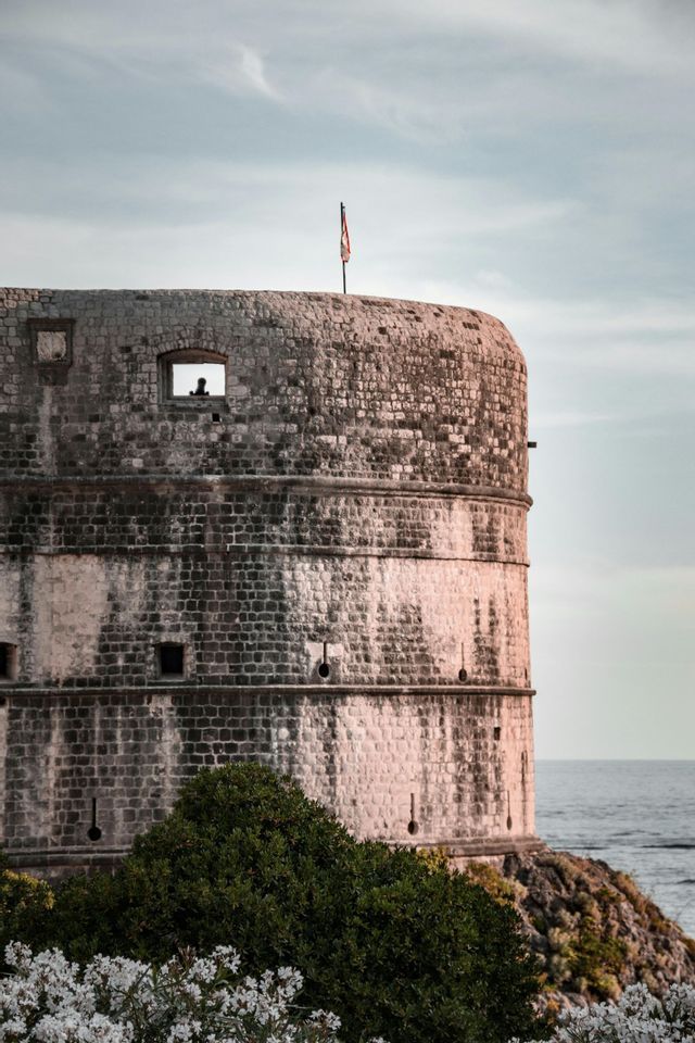 Una torre fortezza in pietra sul mare, con la sagoma di una persona in una finestra e una bandiera sul tetto sotto un cielo nuvoloso.