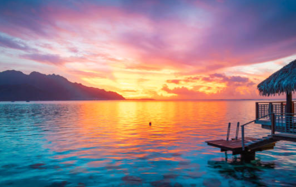 Un bungalow sull'acqua con tetto di paglia si affaccia su un mare calmo sotto un tramonto rosa e arancione vibrante, con montagne in lontananza.