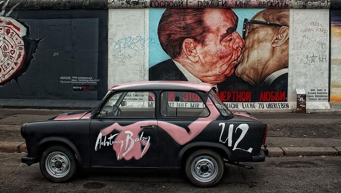 Un coche Trabant negro con grafitis de 'Achtung Baby' está estacionado frente al famoso mural 'Beso Fraternal' en el Muro de Berlín.