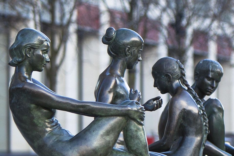 Un primer plano detallado de una escultura de bronce oscuro que representa a cuatro figuras femeninas sentadas juntas en un entorno al aire libre.
