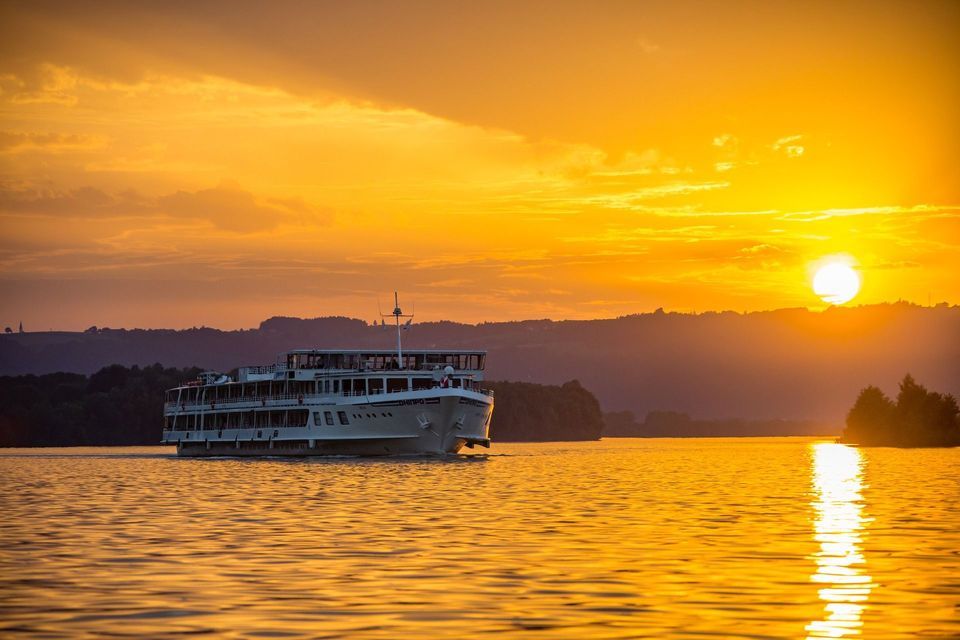 Un crucero blanco navega en aguas tranquilas durante una puesta de sol dorada, con colinas silueteadas al fondo.