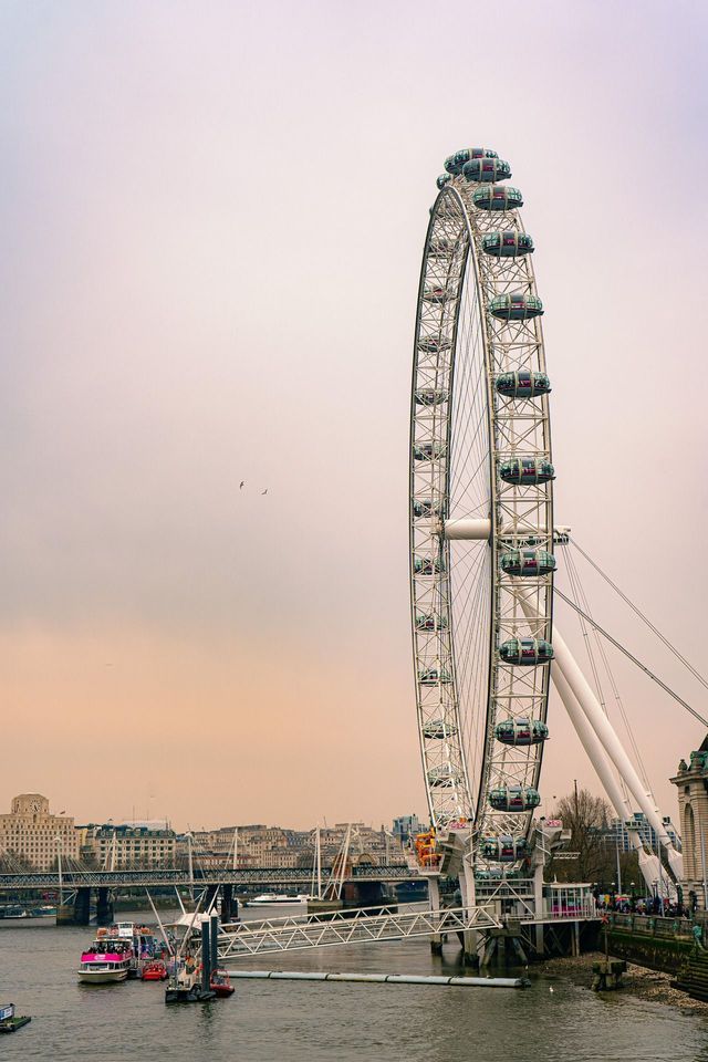 Una gran noria blanca se alza a orillas de un río con el horizonte de la ciudad y un puente de fondo bajo un cielo cubierto.