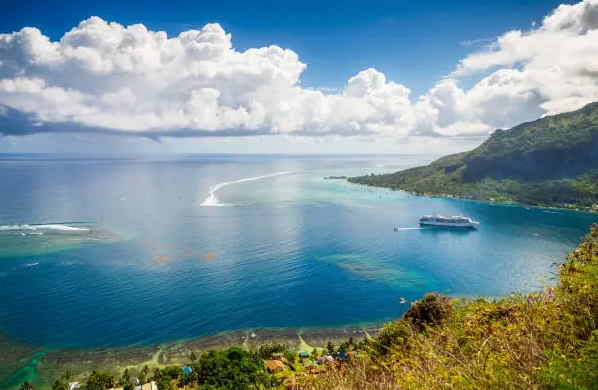 Una vista aerea di una nave da crociera bianca che naviga in una baia turchese accanto a una montagna verde lussureggiante sotto un cielo con grandi nuvole bianche.