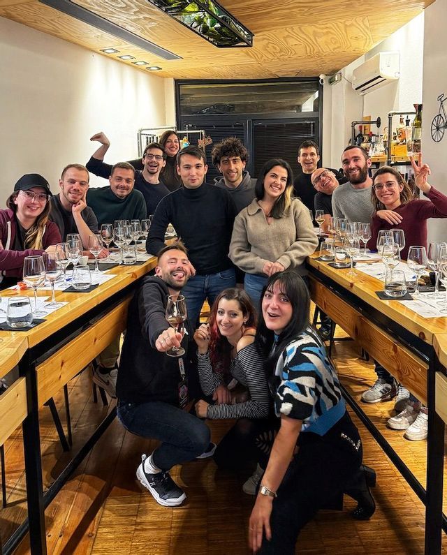 Un gruppo WeRoad posa per una foto intorno a lunghi tavoli di legno apparecchiati con bicchieri da vino durante un evento di degustazione al chiuso.