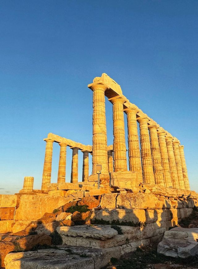 La luce dorata del sole illumina le colonne in pietra di un'antica rovina di tempio contro un cielo azzurro e sereno.