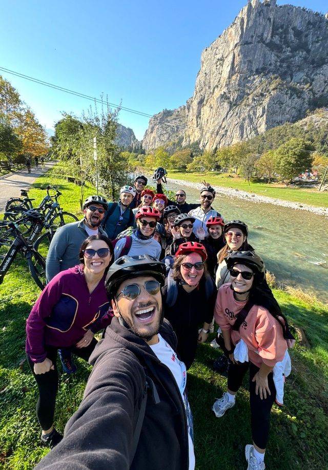 Un gruppo WeRoad in viaggio, con caschi da ciclista, si scatta un selfie su una sponda erbosa del fiume, con ripide montagne sullo sfondo.