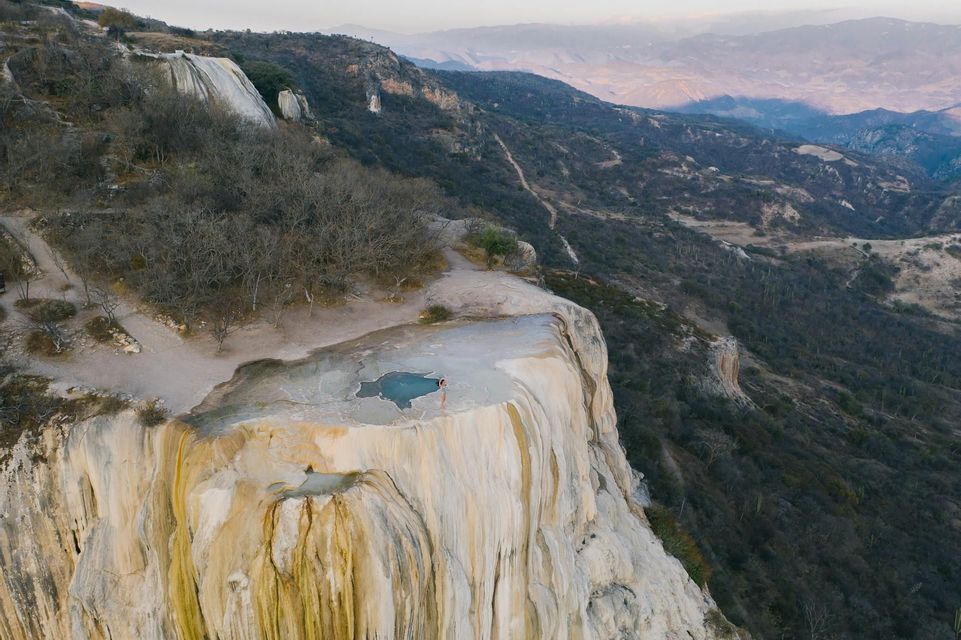 Vista aerea di una persona presso una piscina a sfioro turchese su una cascata pietrificata, affacciata su una valle montuosa.