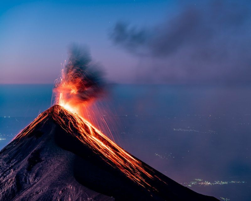 Un vulcano attivo erutta al tramonto, sprigionando lava incandescente e fumo contro un cielo buio, con le luci di città lontane visibili sotto.
