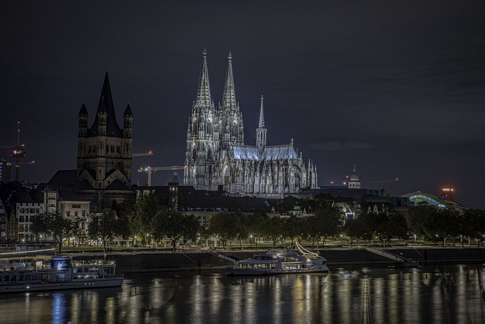 Una cattedrale gotica illuminata con torri gemelle si erge su una città di notte, con un fiume e barche in primo piano.