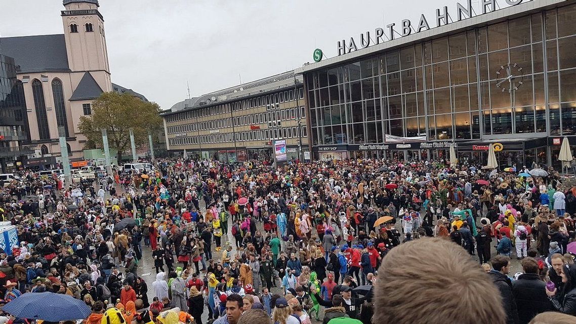 Una grande folla di persone, molte con costumi colorati, riempie una piazza della città di fronte a un edificio moderno con una facciata in vetro.