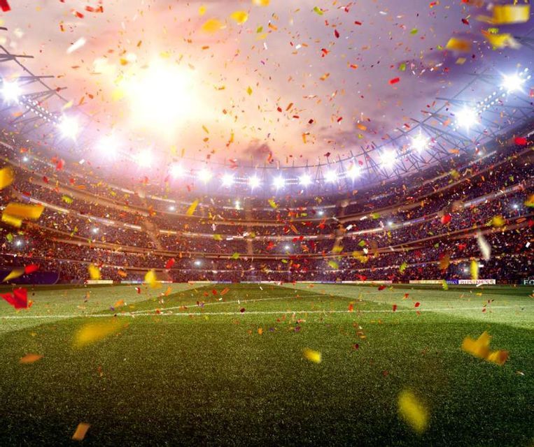 Colorful confetti falls onto the green grass field of a packed sports stadium illuminated by bright floodlights.
