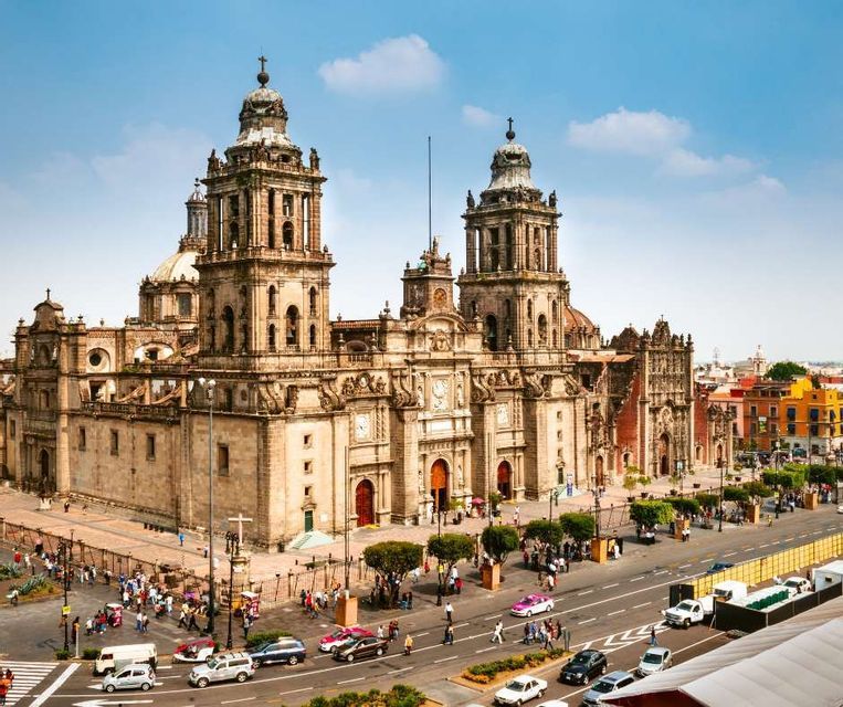 An aerial view of a large, historic cathedral with twin bell towers, next to a busy street and plaza with people and traffic.