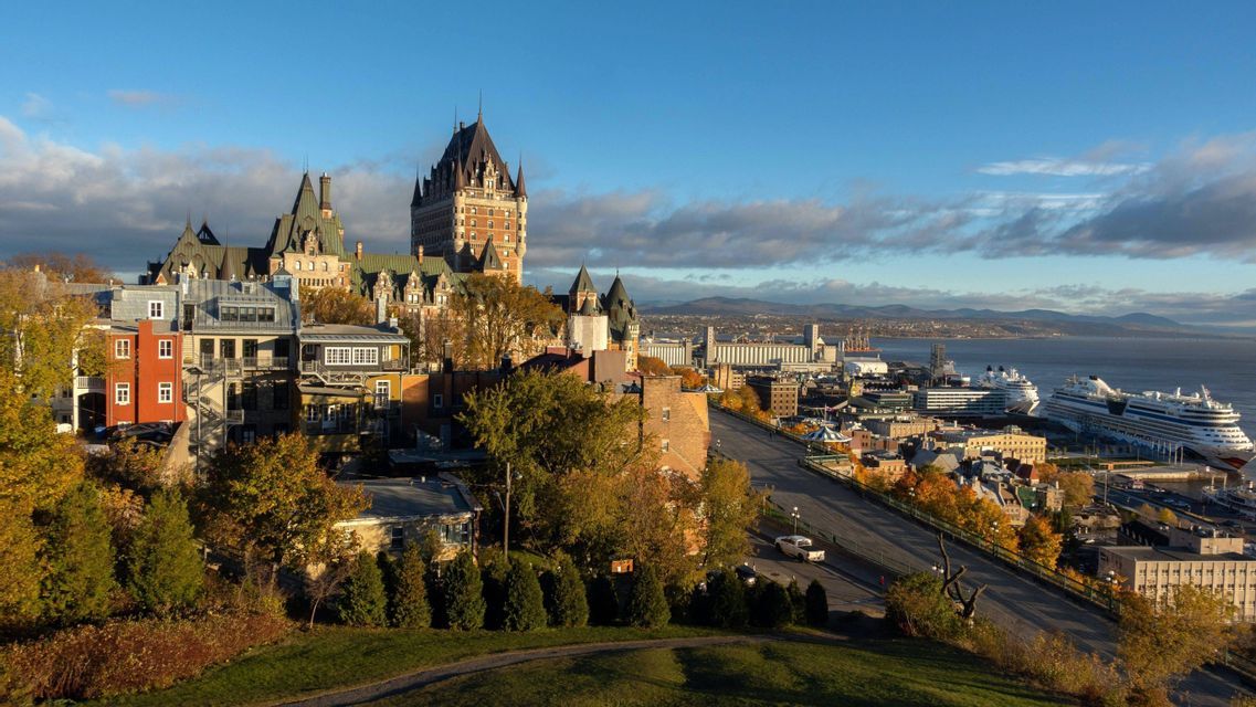 Un paysage urbain avec un bâtiment historique ressemblant à un château, des arbres d'automne et un port fluvial avec des navires de croisière amarrés.