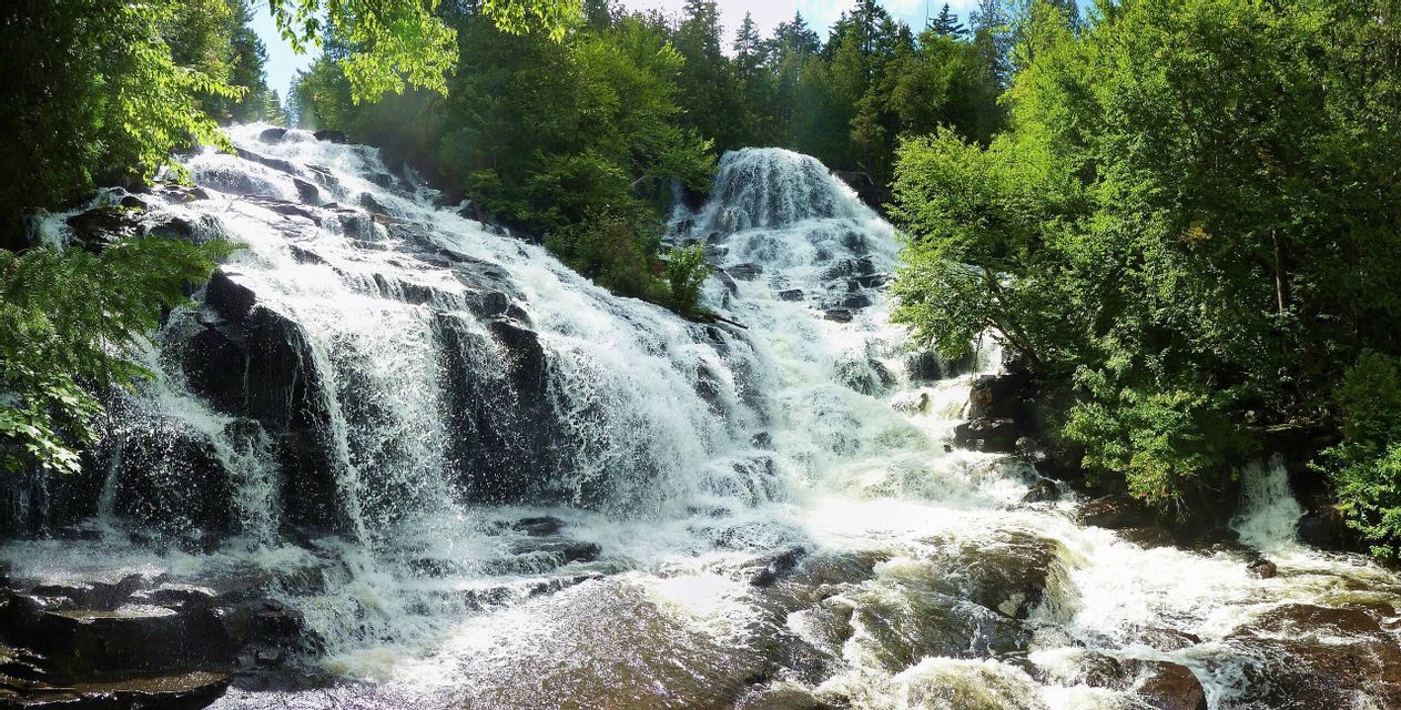 Una cascata ampia e a più livelli scende lungo un pendio roccioso, immersa in una fitta foresta verde sotto un cielo luminoso.