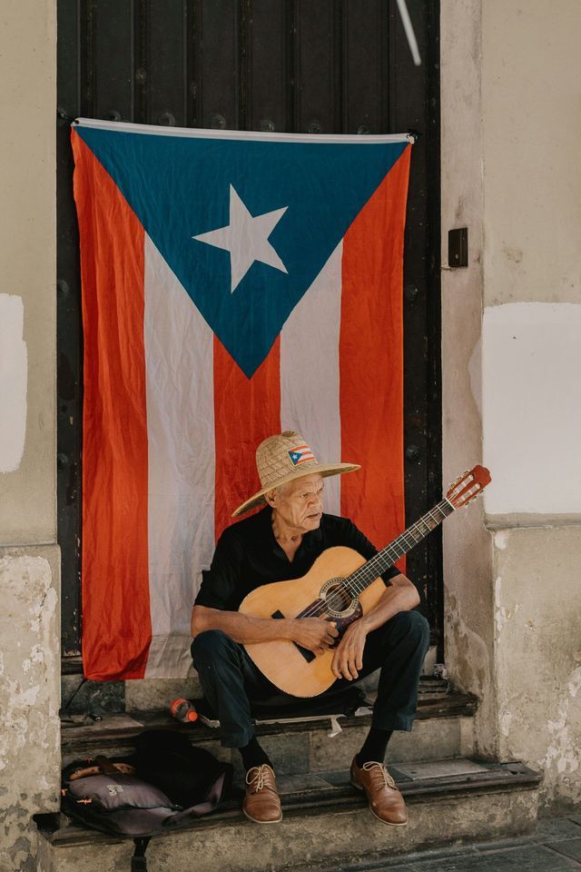 Un anciano con sombrero de paja toca una guitarra sentado en unas escaleras frente a una gran bandera puertorriqueña.