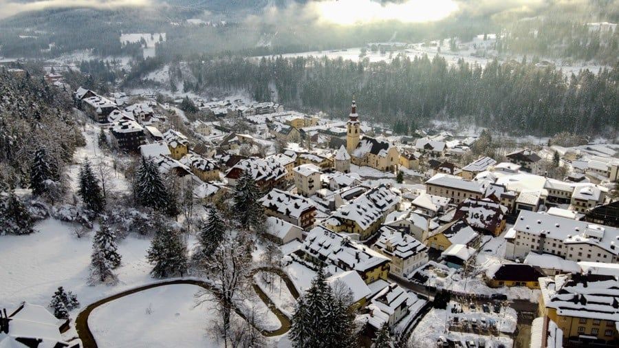 Una vista aerea di un piccolo villaggio di montagna con tetti innevati, circondato da una fitta pineta in inverno.