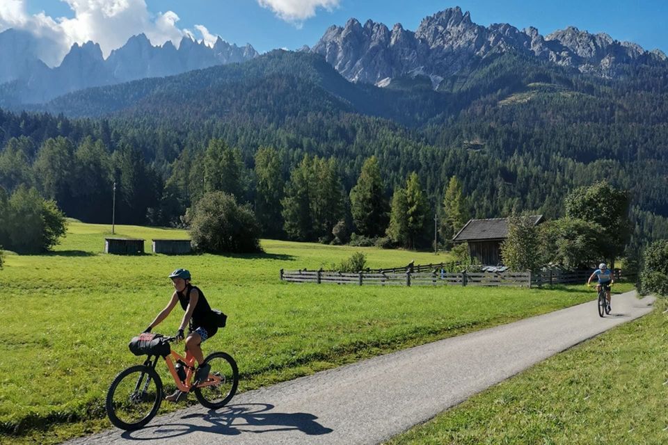 Eine WeRoad Gruppen-Fahrradtour auf einem asphaltierten Weg in einem Gebirgstal, mit einem Wald und zerklüfteten Gipfeln im Hintergrund.