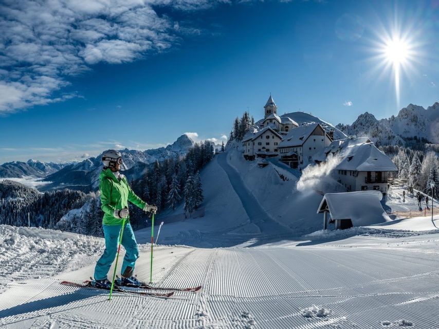 Uno sciatore con la giacca verde si erge su una pista da sci appena battuta, ammirando un paese innevato e le montagne.
