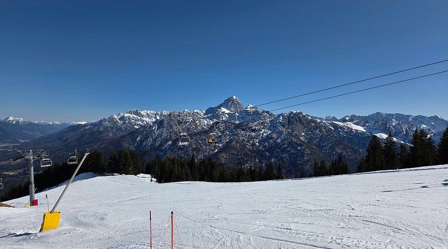 Una vista da una pista da sci innevata mostra una seggiovia che sale verso una catena di montagne innevate sotto un cielo azzurro e limpido.