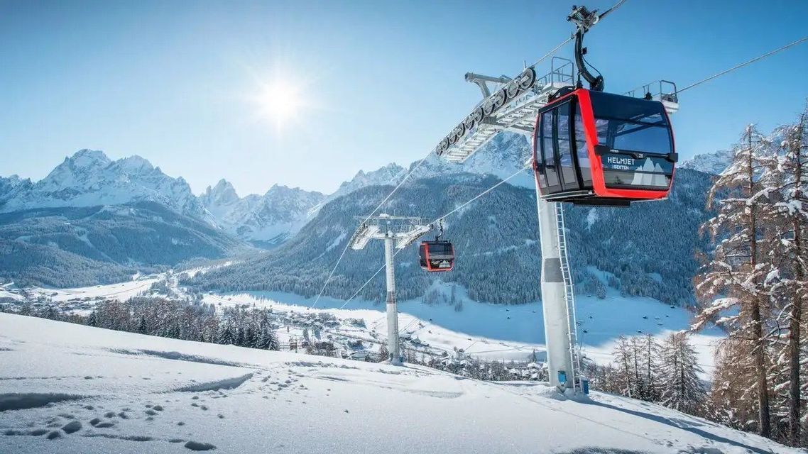 Teleféricos rojos y negros viajan sobre una pendiente de montaña nevada en un día soleado, con una vasta cordillera al fondo.