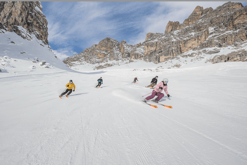 Un viaje en grupo de cinco personas de WeRoad esquiando por una amplia pista nevada rodeada de imponentes montañas rocosas bajo un cielo azul.