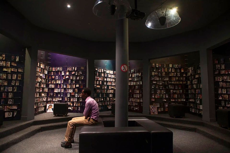 Un homme est assis sur un banc dans une salle commémorative circulaire faiblement éclairée, contemplant les murs recouverts de rangées de portraits photographiques.