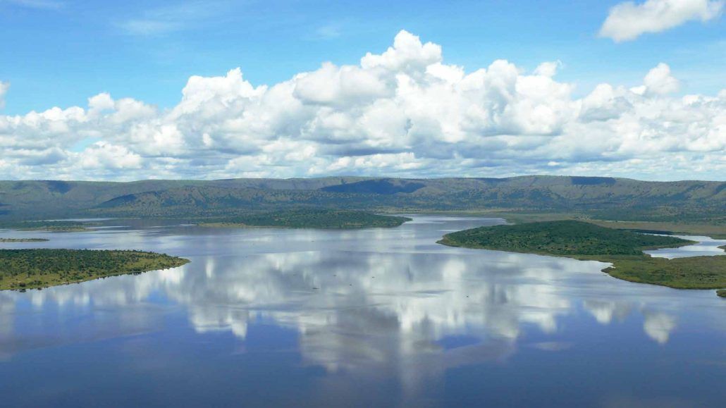 Un vaste lac calme reflète un ciel bleu avec des nuages blancs et cotonneux, entouré de vertes collines ondulantes en arrière-plan.