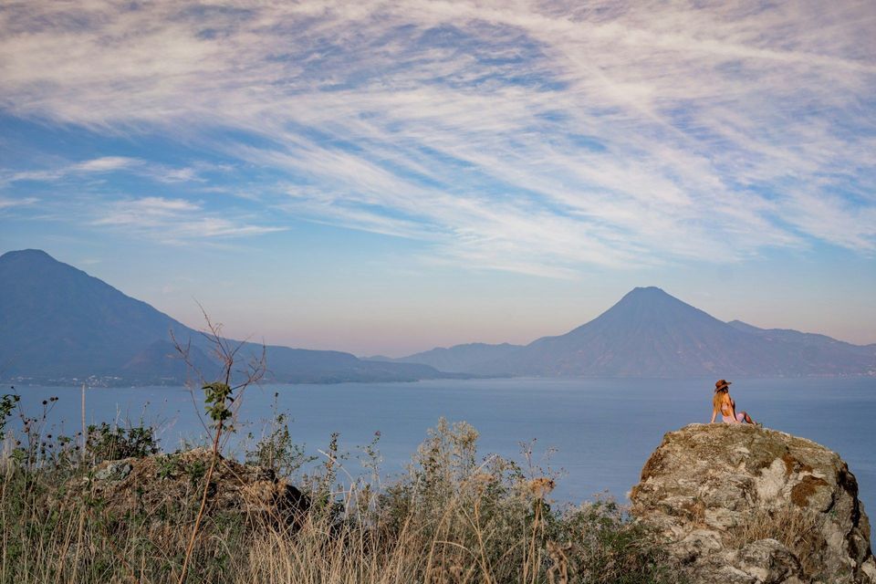 Una persona con un cappello è seduta su una grande roccia, ammirando un vasto lago circondato da montagne sotto un cielo nuvoloso.