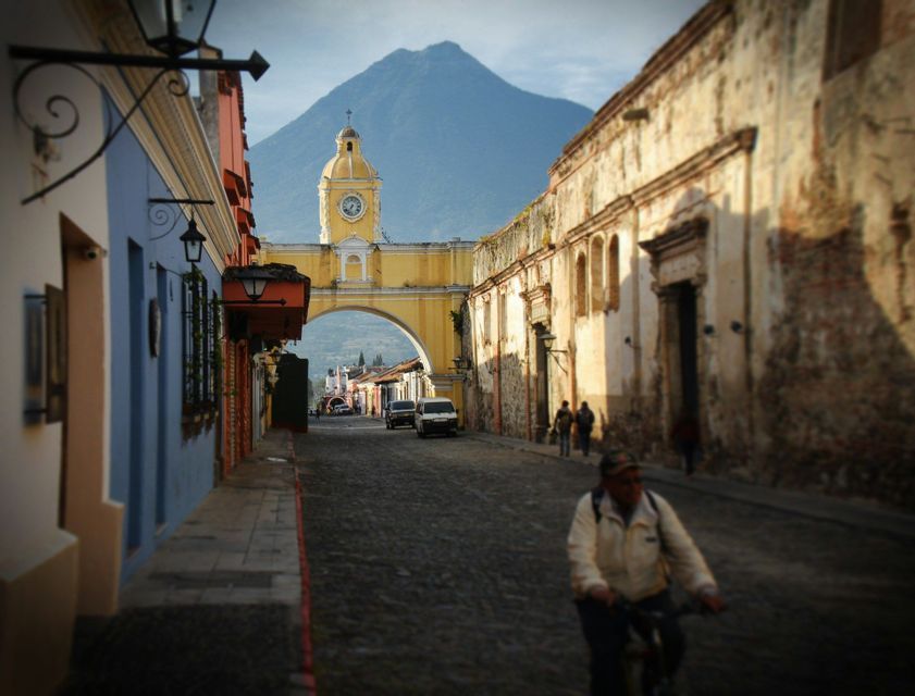 Una persona in bicicletta percorre una strada acciottolata verso un arco giallo con una torre dell'orologio, con un grande vulcano sullo sfondo.