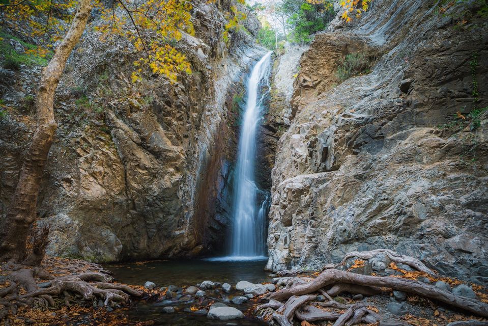 Une fine cascade coule le long d'une falaise rocheuse escarpée dans une petite mare en contrebas, encadrée par un arbre aux feuilles d'automne jaunes.