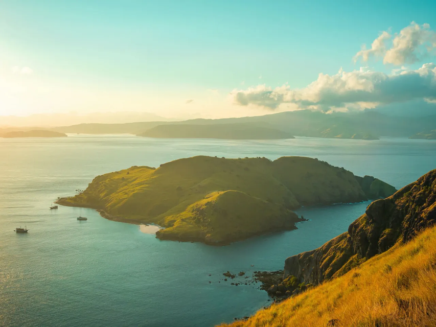 Vista aerea di isole verdi e collinari in acque calme e azzurre, con alcune barche ancorate al largo durante un tramonto dorato.