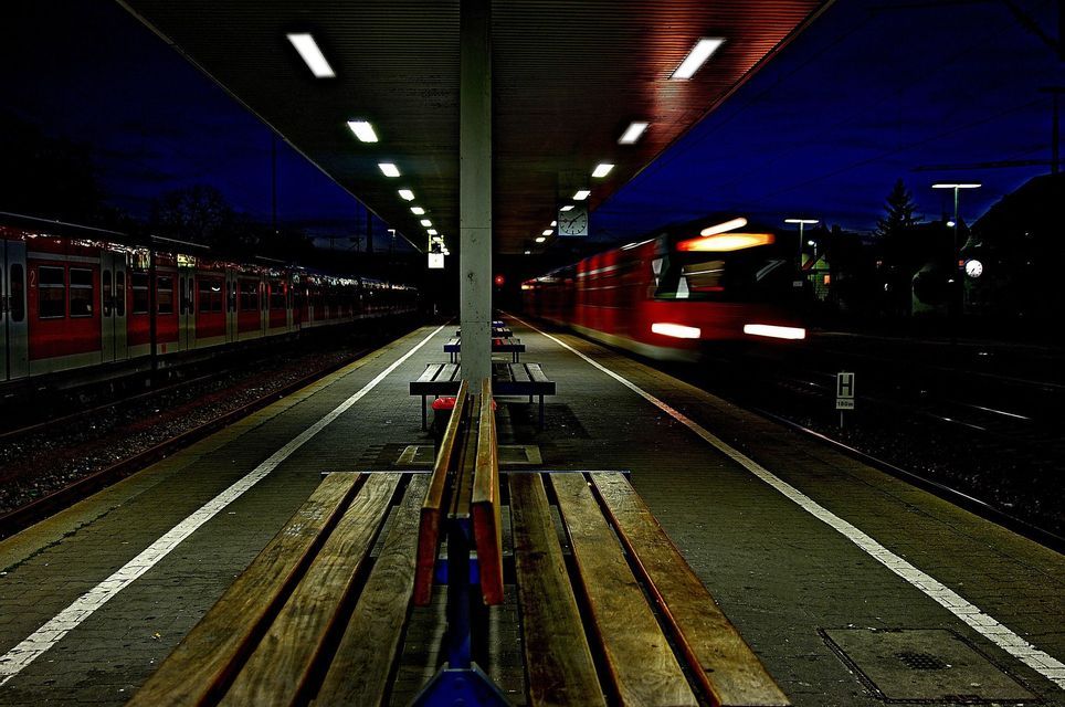 Un treno rosso crea un effetto mosso mentre sfreccia di notte davanti a una banchina della stazione vuota e illuminata, con un altro treno fermo di fronte.