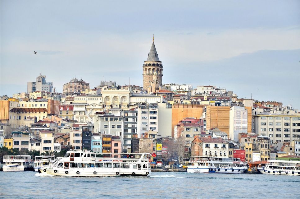 Un traghetto bianco naviga sull'acqua con la Torre di Galata e un denso skyline cittadino sullo sfondo sotto un cielo pallido.