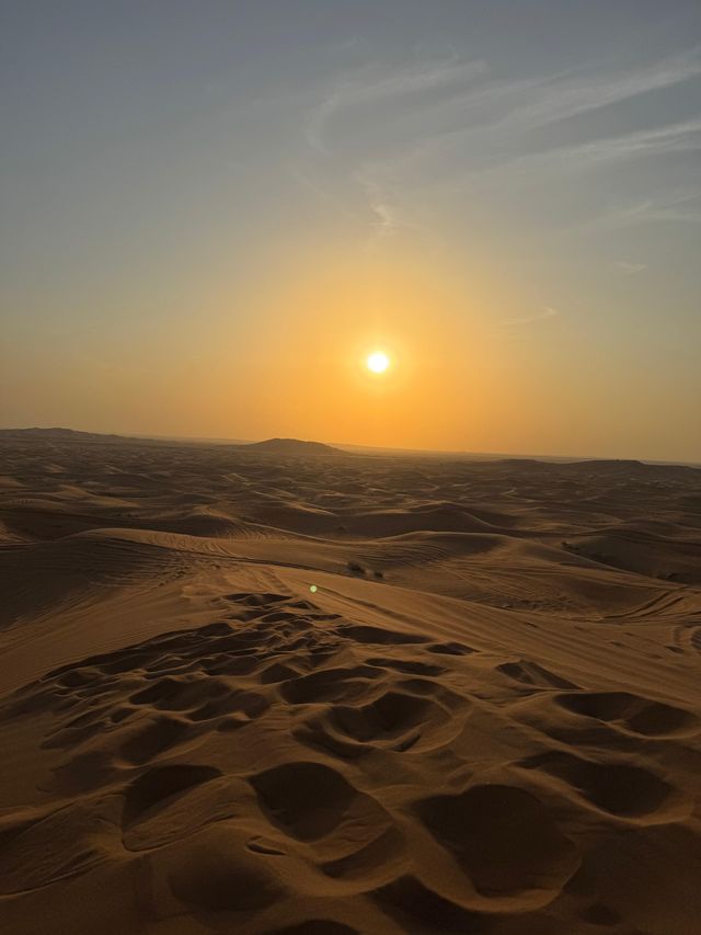 Il sole al tramonto proietta un bagliore dorato su una vasta distesa di sinuose dune di sabbia desertiche sotto un cielo velato.