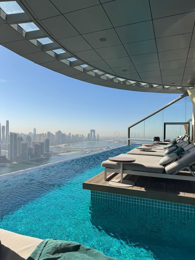 Piscina a sfioro panoramica sul tetto con lettini vuoti su una terrazza in legno, che si affaccia su uno skyline moderno e il mare sotto un cielo sereno.
