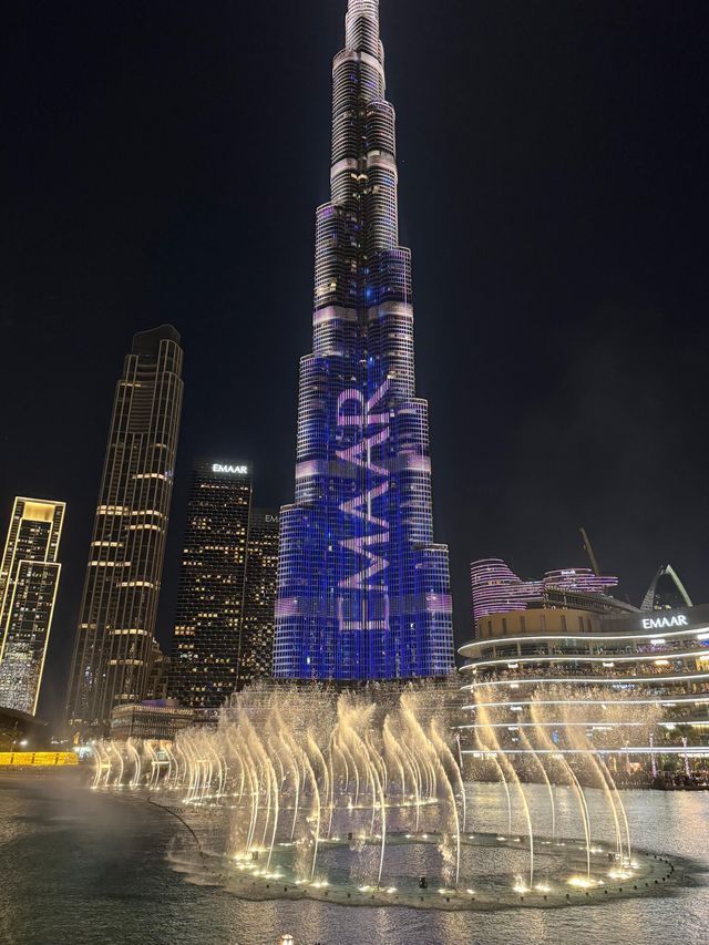 Un grattacielo illuminato con un logo blu si erge di notte dietro una fontana danzante e illuminata in una città.