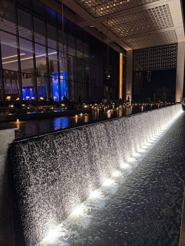 Una cascata artificiale, illuminata dal basso, scende lungo una parete accanto a un moderno edificio con facciata in vetro, di notte.