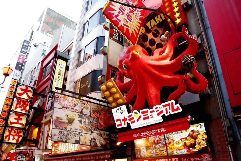 Una grande scultura di polpo rosso su una facciata di edificio, circondata da molti cartelli colorati con caratteri giapponesi su una strada cittadina.