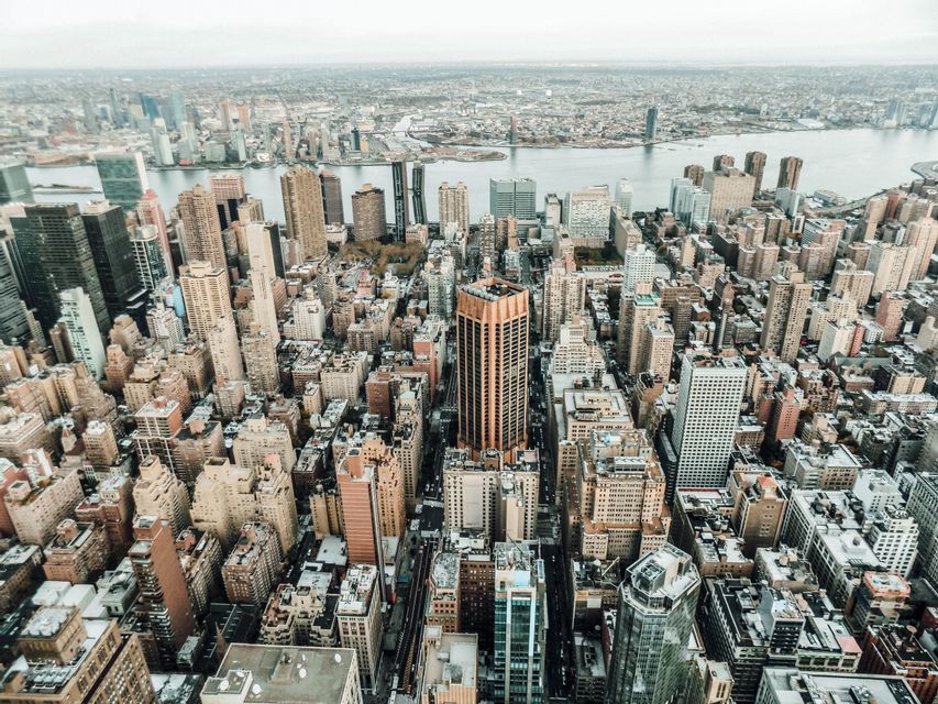 Luftaufnahme einer dichten Stadtlandschaft mit Wolkenkratzern und einem breiten Fluss im Hintergrund an einem bewölkten Tag.