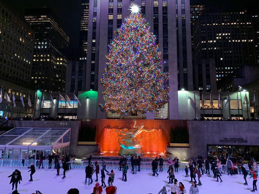 Der Weihnachtsbaum des Rockefeller Centers, hell erleuchtet mit bunten Lichtern, überragt nachts die Menschen, die auf der Eisbahn darunter Schlittschuh laufen.