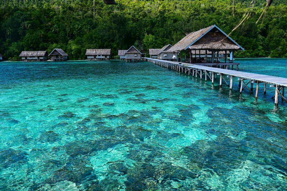 Un lungo pontile in legno collega bungalow sull'acqua sopra un mare turchese cristallino, con un'isola verde e lussureggiante sullo sfondo.