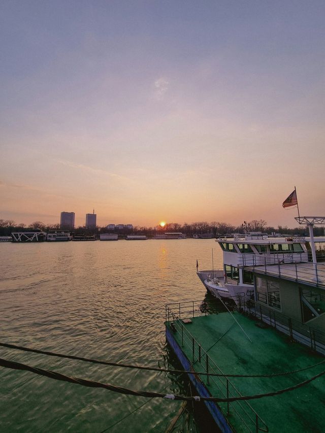 Il sole tramonta su un ampio fiume con barche ormeggiate lungo la riva e uno skyline della città visibile in lontananza.