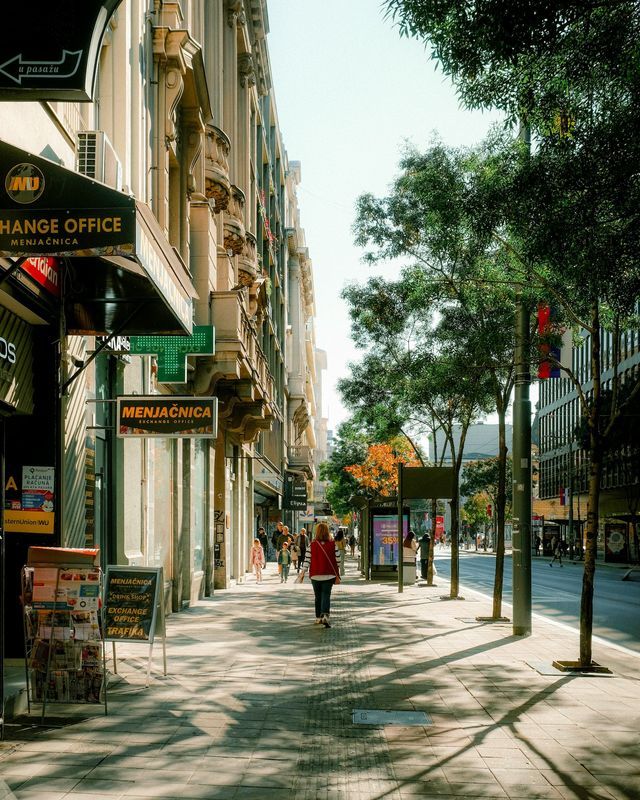 Una donna con un gilet rosso cammina lungo un marciapiede cittadino illuminato dal sole, costeggiato da edifici decorati e alberi frondosi.