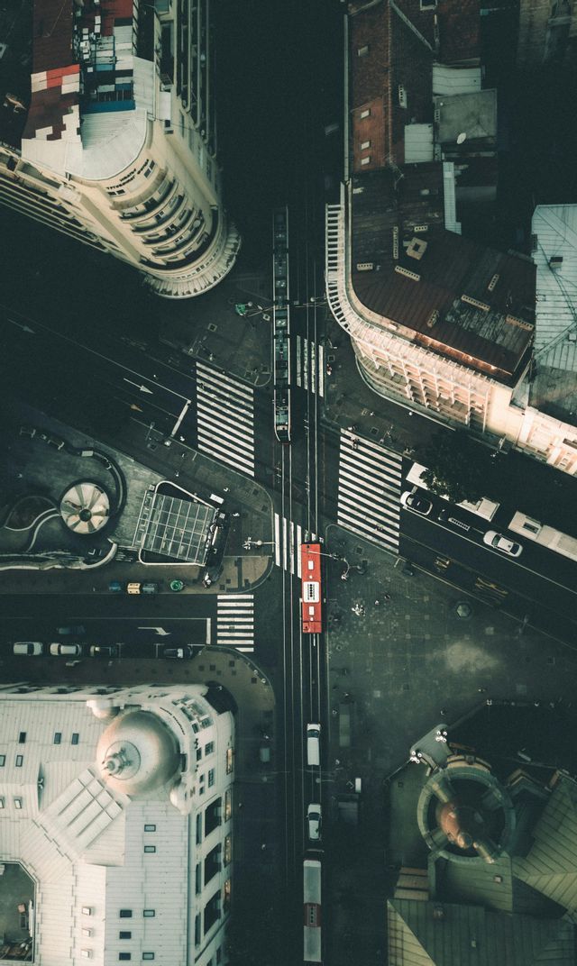 Veduta aerea dall'alto di un incrocio urbano con un tram rosso sui binari, auto e strisce pedonali tra palazzi alti.